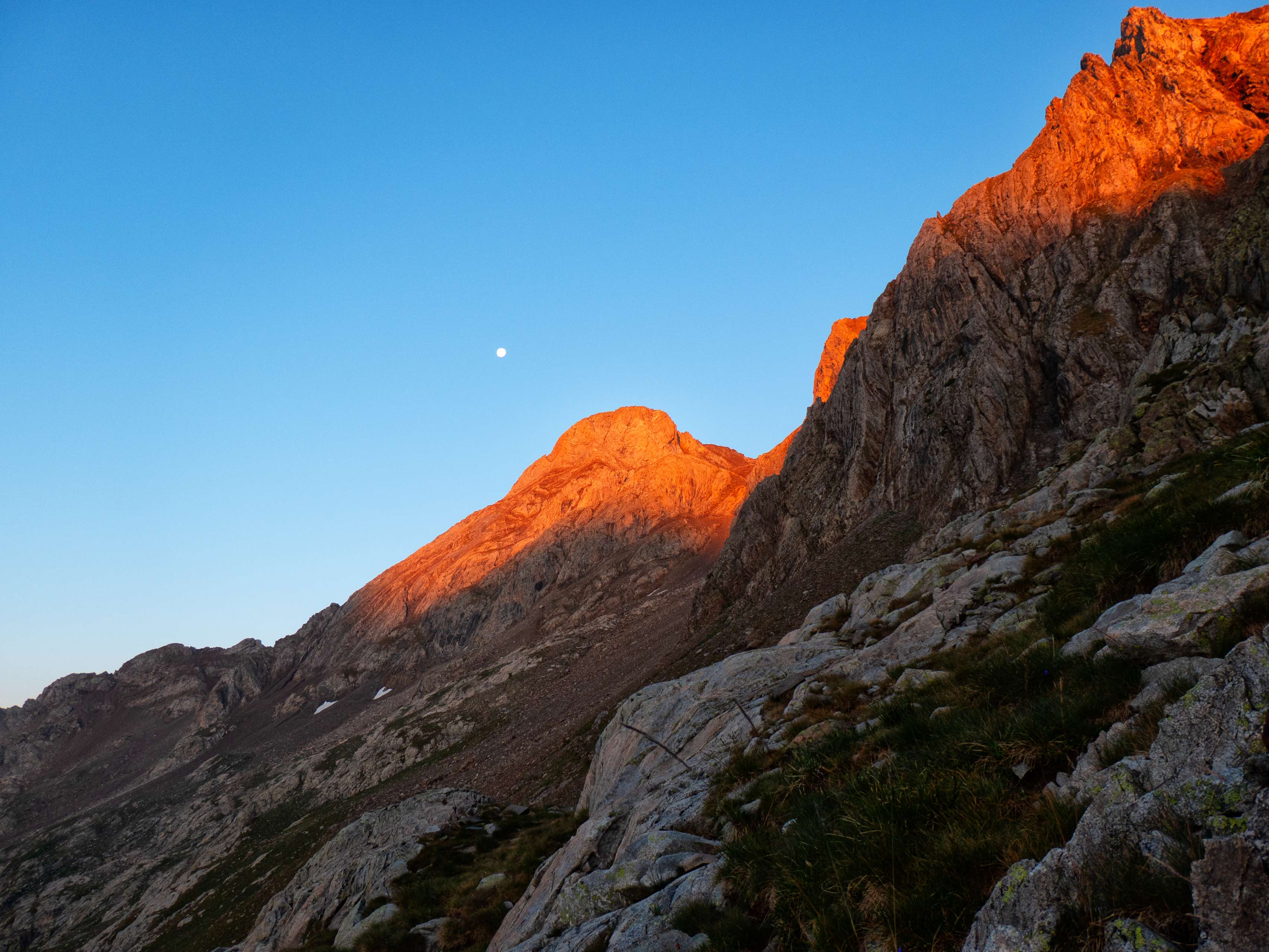 El rosado da paso al rojo, aún más intenso cuando se refleja en la colorida roca metamórfica predominante en los picos de las Argualas, aquí con la cumbre homónima acompañada por la luna. La ruta normal a casi todas las cimas del macizo discurre por la pedrera situada a los pies del Garmo Negro, por la que el sendero que vemos en la parte inferior de la foto traza una larga diagonal buscando el collado de las Argualas, que da acceso al circo somital.